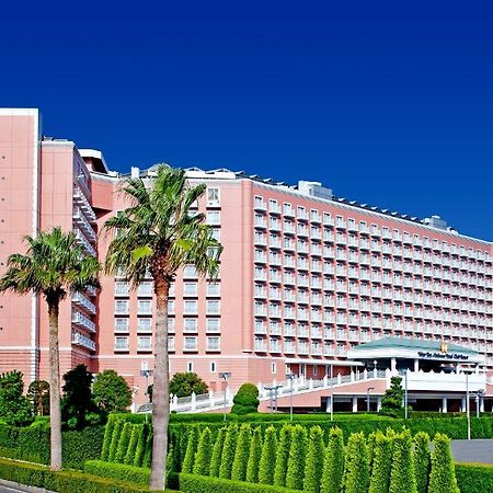 Grand Nikko Tokyo Bay Maihama Hotel Urajaszu Kültér fotó The image shows a large, multi-story hotel building featuring a light pink facade. The architecture appears modern, with numerous windows arranged in a grid pattern. In the foreground, there are neatly trimmed shrubs and palm trees, adding a tropical