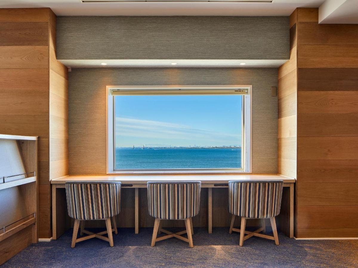 Grand Nikko Tokyo Bay Maihama Hotel Urajaszu Kültér fotó The photo shows a modern workspace with three striped chairs positioned at a long, white desk. The desk faces a large window with a view of a tranquil body of water and a distant skyline. The walls around the workspace are made of wood paneling, crea