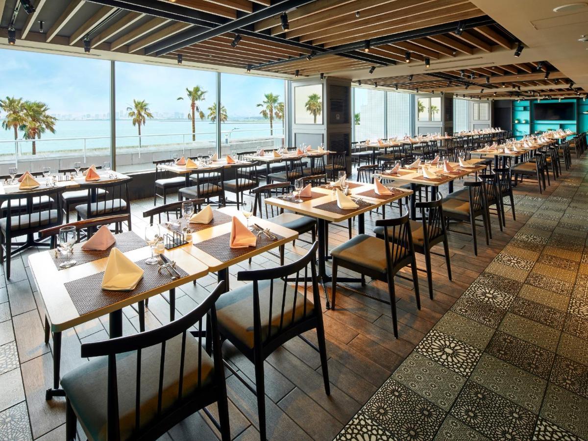 Grand Nikko Tokyo Bay Maihama Hotel Urajaszu Kültér fotó The photo shows a modern restaurant interior featuring rows of neatly arranged tables and chairs. The large windows provide a scenic view of palm trees and what appears to be a body of water in the background, suggesting a coastal location. Each tabl