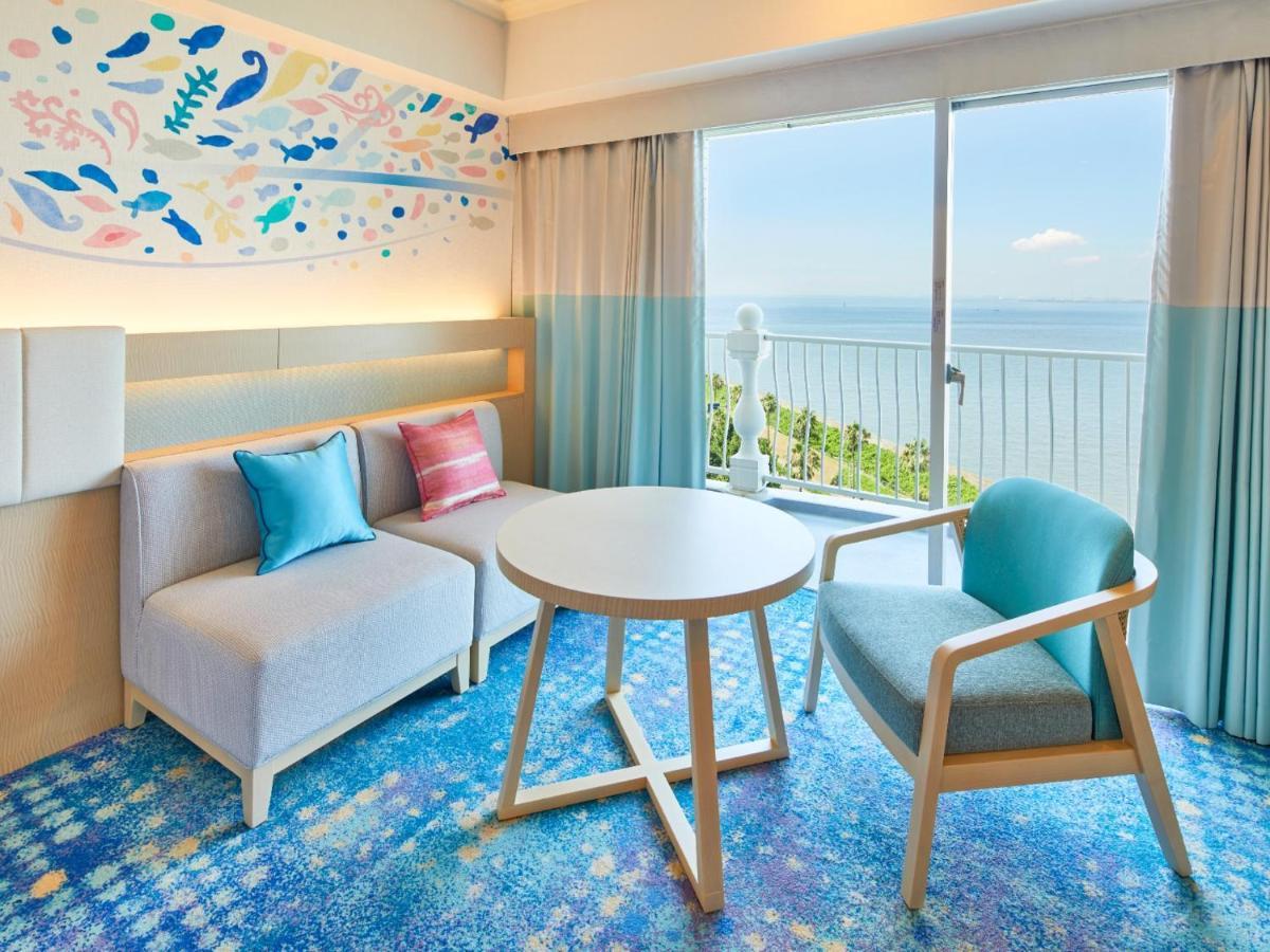 Grand Nikko Tokyo Bay Maihama Hotel Urajaszu Kültér fotó The photo shows a modern and stylish interior of a room with a view. There is a cozy seating area featuring a light-colored sofa with decorative cushions in various shades of blue and pink. A round table occupies the center of the space, surrounded b