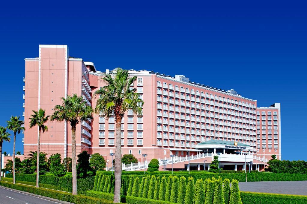 Grand Nikko Tokyo Bay Maihama Hotel Urajaszu Kültér fotó The image shows a large, multi-story hotel building featuring a light pink facade. The architecture appears modern, with numerous windows arranged in a grid pattern. In the foreground, there are neatly trimmed shrubs and palm trees, adding a tropical